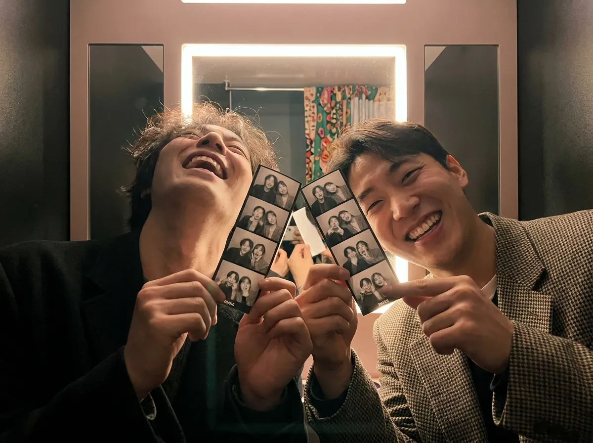 Hongdae Photo Booth Experience Seoul inside booth with two friends laughing and reviewing printed photo strips under warm light