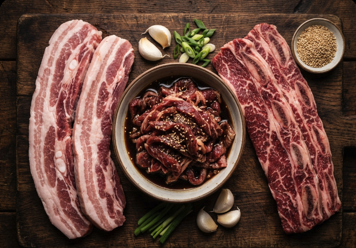Korean BBQ at home for beginners meat cuts: Samgyeopsal pork belly, bulgogi, and galbi short ribs on a wooden board with garlic.