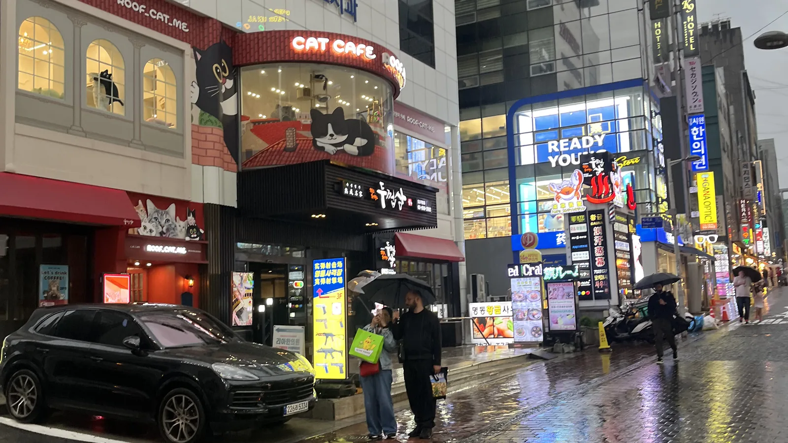 Myeongdong at night view of a rainy street corner with a glowing cat cafe and a few umbrellas
