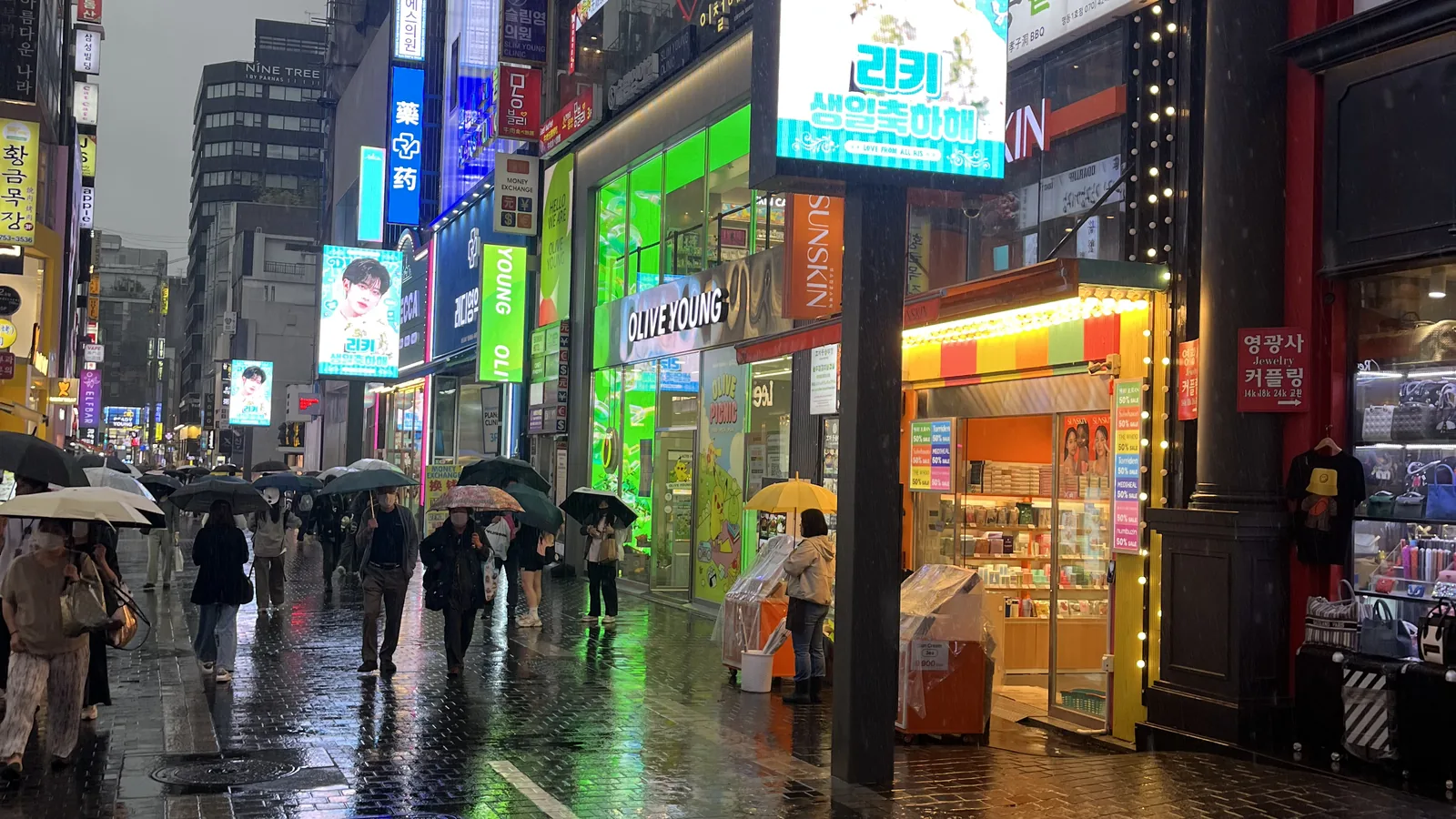 Myeongdong at night in the rain with neon signs reflecting on the wet street and a row of umbrellas